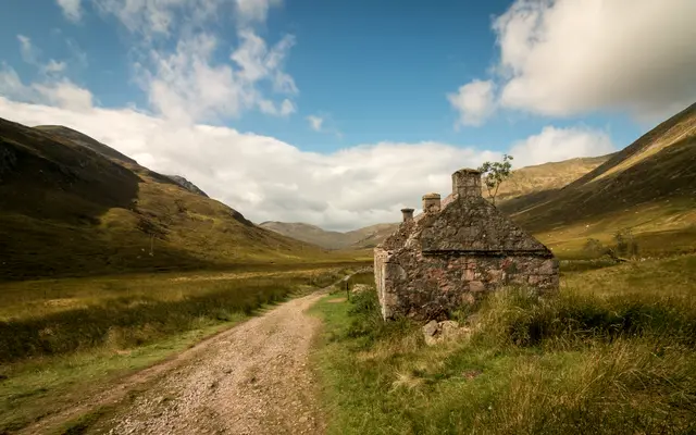 Dirt road winding through a green valley past a stone cottage